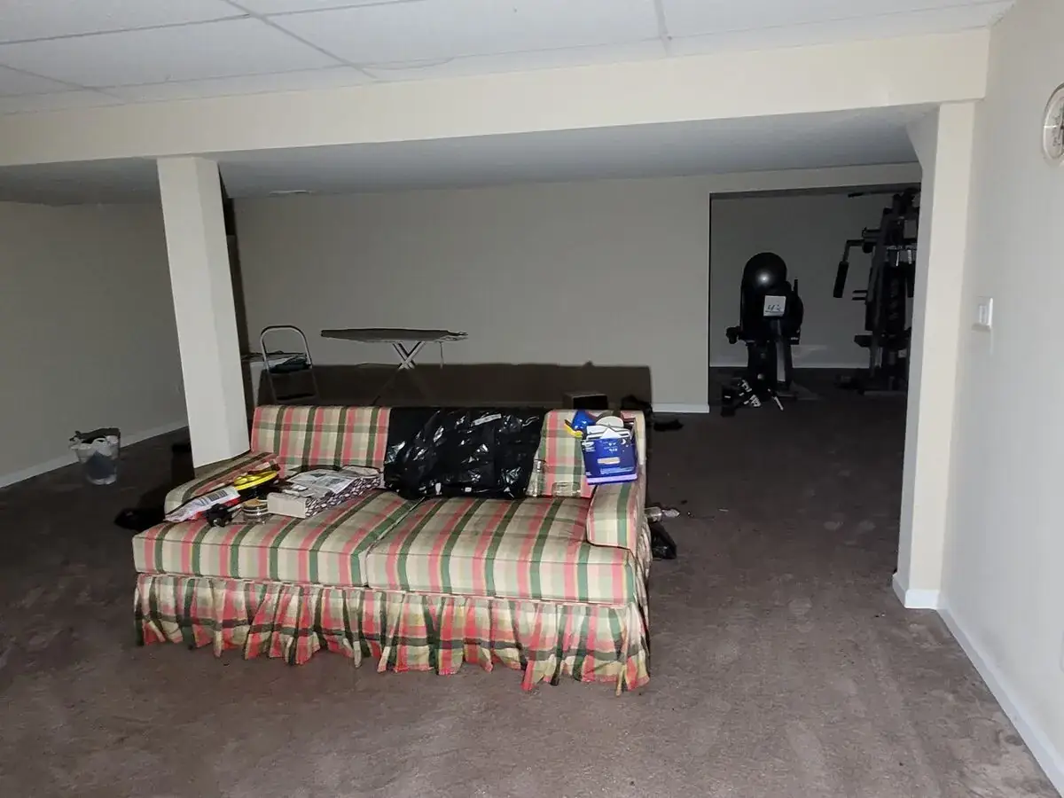 Basement living area before cleanup, with furniture and belongings remaining on the carpet.